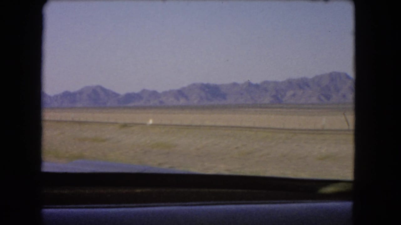1966 Nevada road footage shows an arid plain, distant mountains, and a small white roadside marker through the car window.
