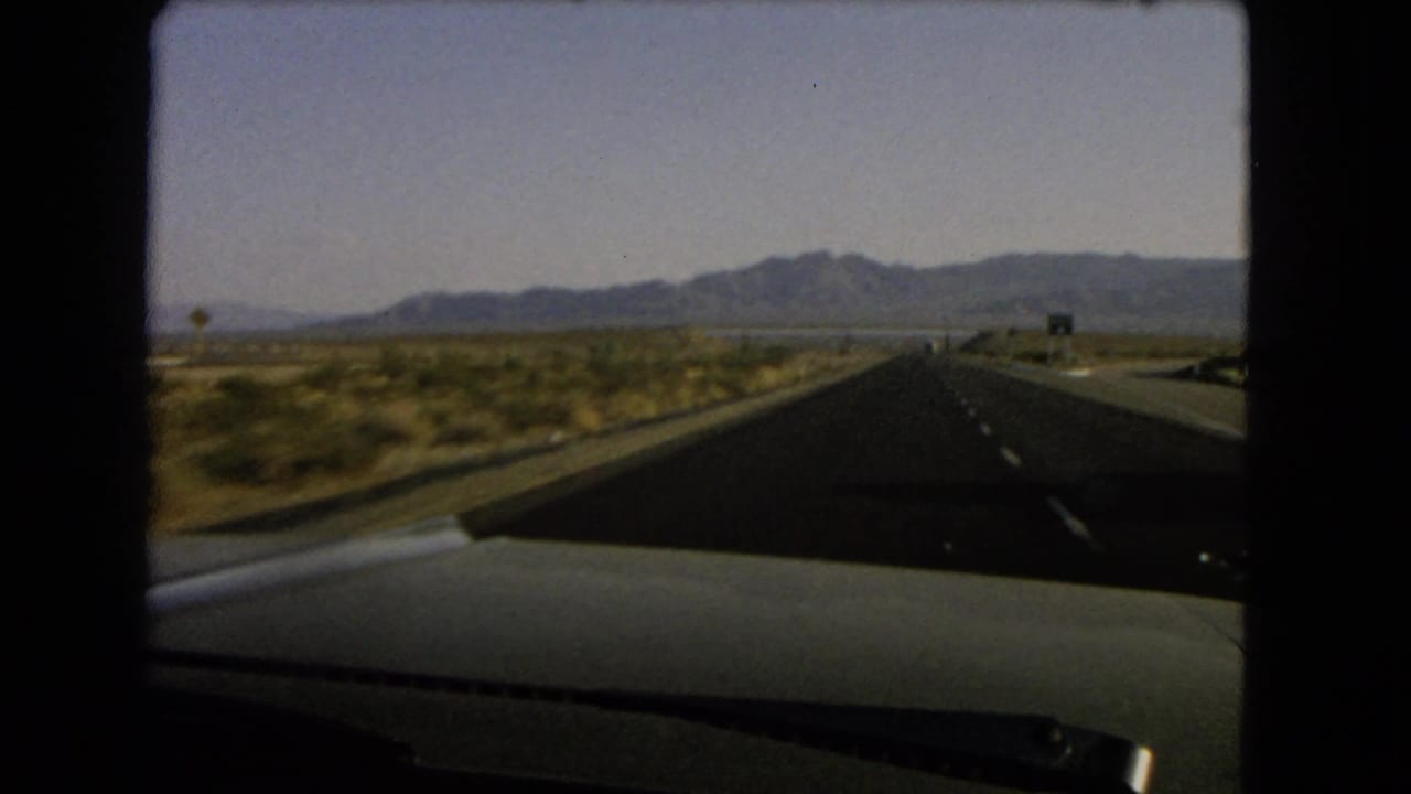 1966 Nevada highway view from inside a moving car, showing a straight road through desert scrub toward low mountains.
