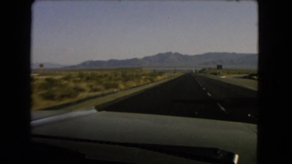 1966 Nevada highway view from inside a moving car, showing a straight road through desert scrub toward low mountains.