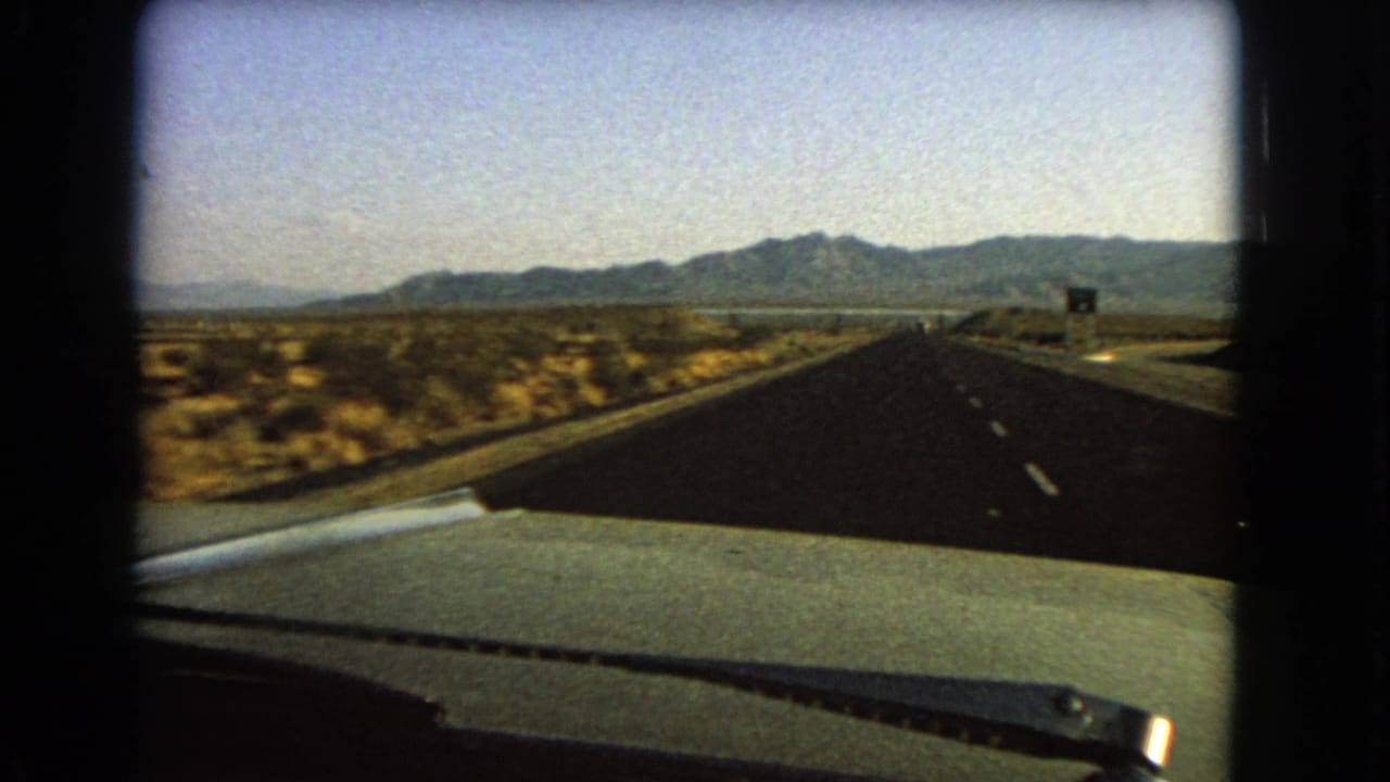 Windshield perspective on a Nevada desert highway in 1966, with sparse brush, mountains, and roadside signage ahead.
