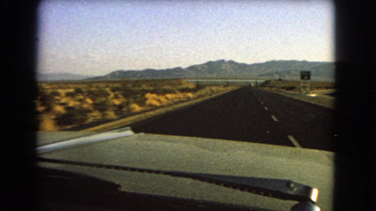 1966 Nevada road-trip frame from inside a car, looking over the dashboard toward a long desert highway and mountains.
