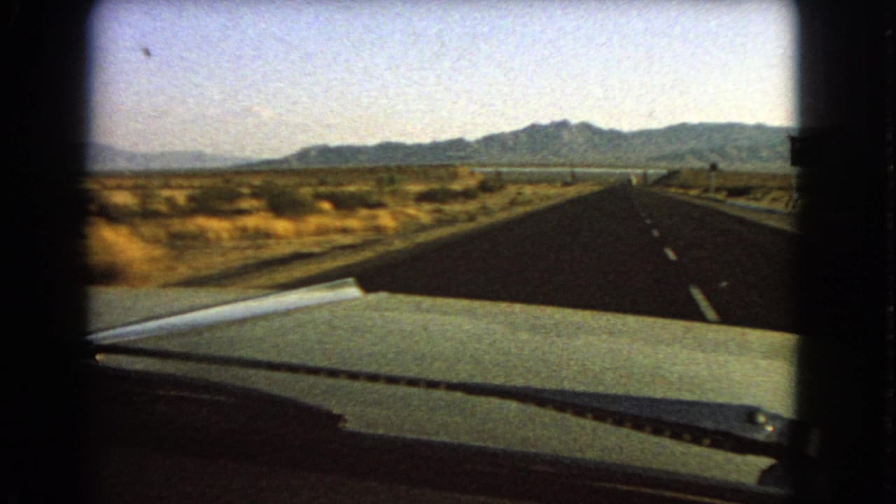 Windshield view on a 1966 Nevada desert highway, showing road markings, scrubland, signage, and low mountains ahead.