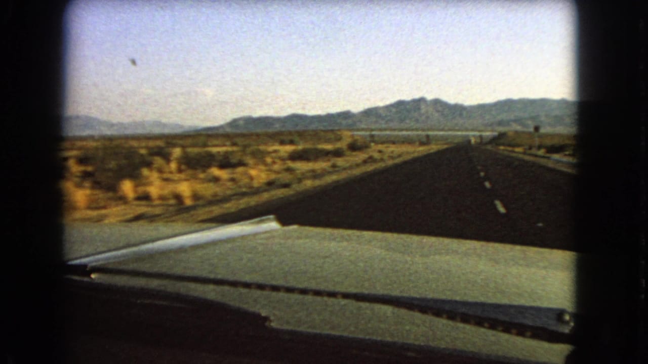 1966 Nevada highway seen from inside a car, with the hood and windshield wiper in front of desert brush and distant mountains.