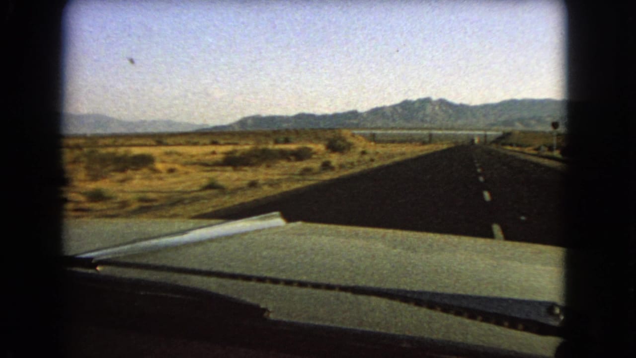 Road through arid terrain with car hood and mountain range ahead.