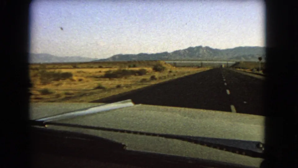 Car interior viewpoint on a Nevada desert road, showing the hood, wiper, empty highway, low brush, and mountains in the distance.