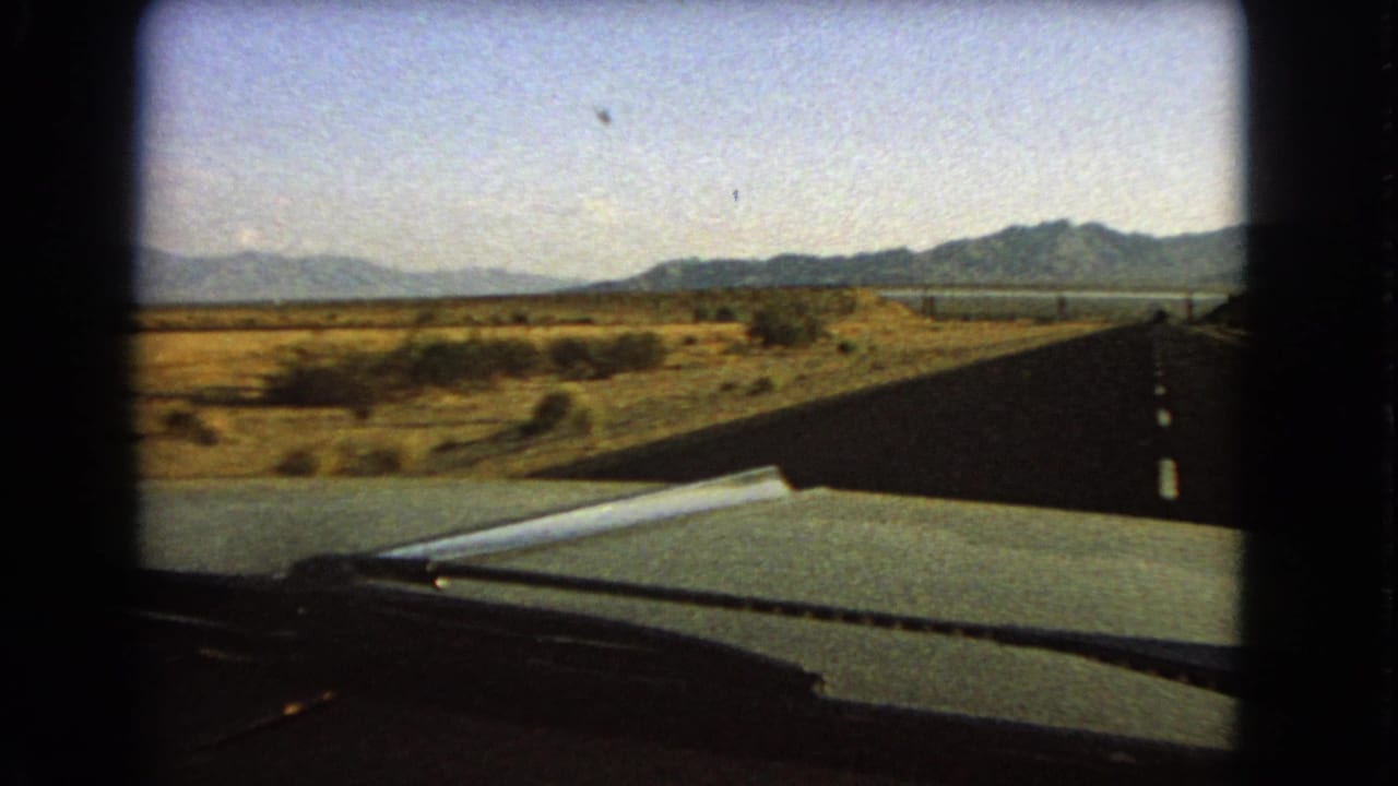 1966 car-window view across a Nevada highway, with desert scrub beside the road and mountains visible beyond the windshield.