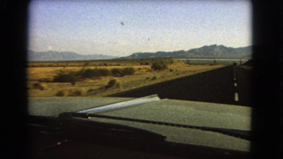 1966 car-window view across a Nevada highway, with desert scrub beside the road and mountains visible beyond the windshield.