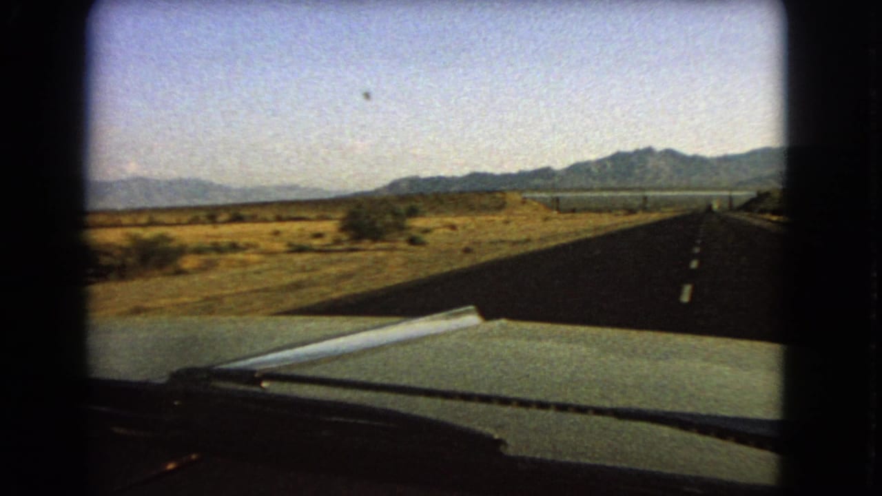 Nevada road-travel frame from inside a car, showing the hood, wiper, straight highway, roadside desert, and distant mountain ridge.