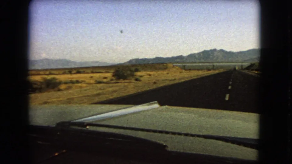 Nevada road-travel frame from inside a car, showing the hood, wiper, straight highway, roadside desert, and distant mountain ridge.