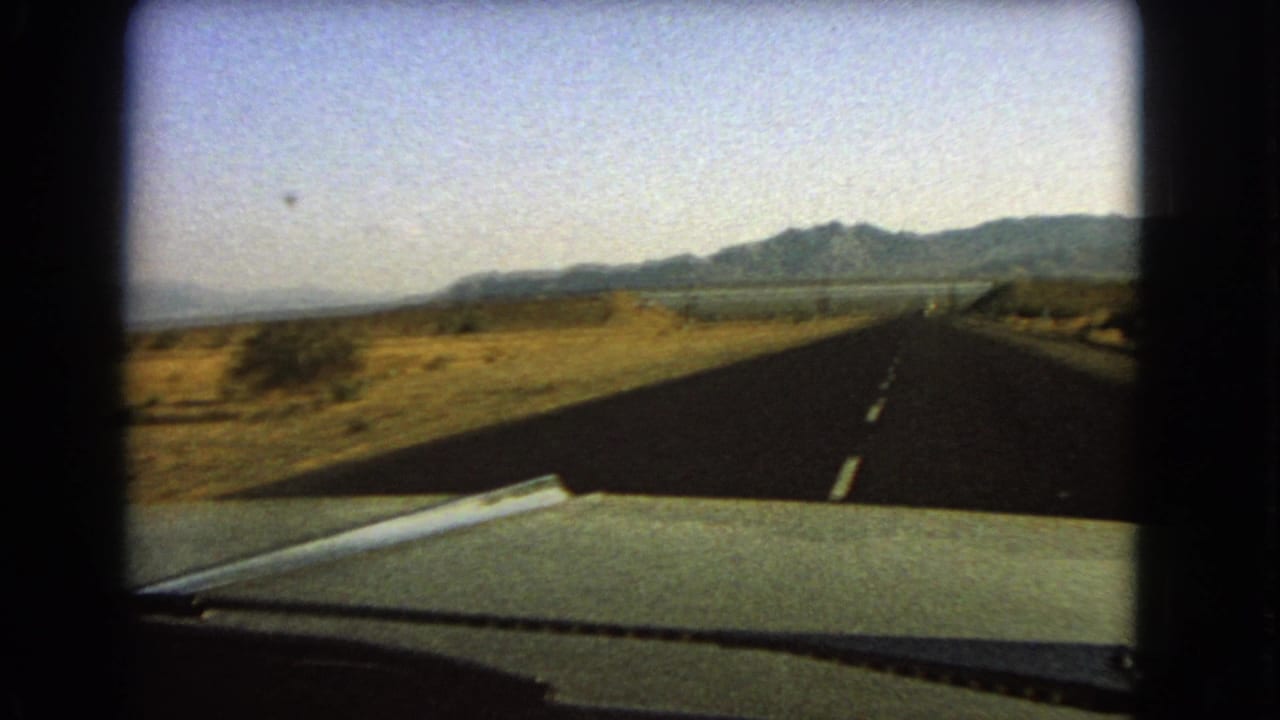 Straight desert highway seen over a car hood with mountains in the distance.