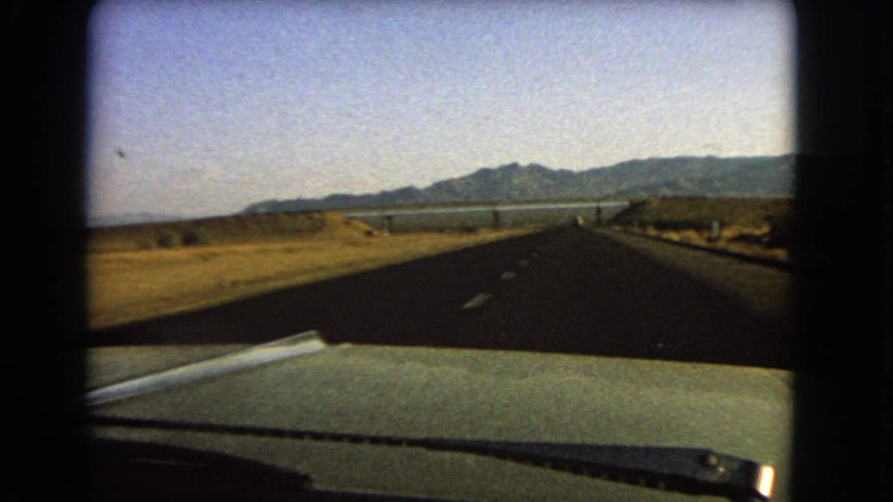 A 1966 Nevada home-movie frame shows a moving car view along a highway through dry terrain toward water and mountains.