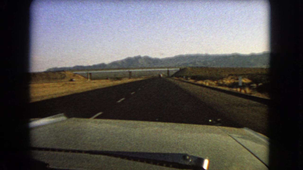 Final 1966 Nevada frame from inside a car, showing a straight desert highway leading toward distant water and mountains.