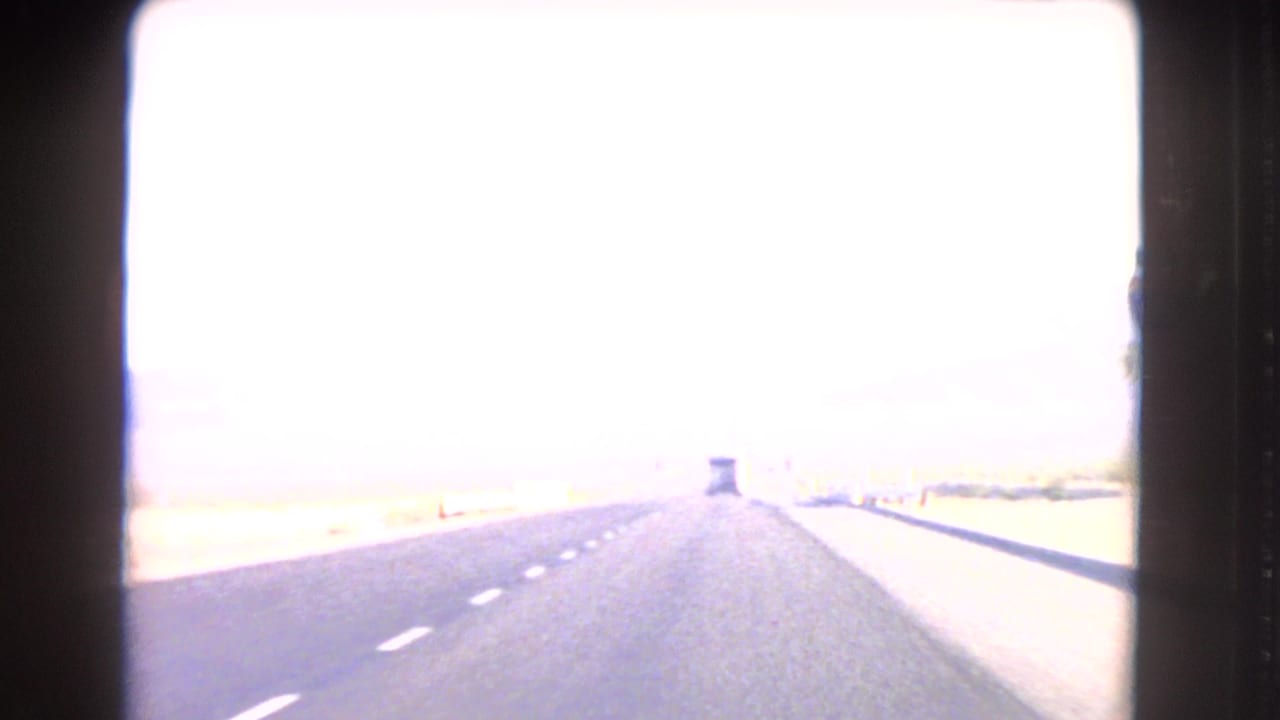 1966 8mm POV from a car on a Nevada desert highway, with bright exposure, lane markings, and a vehicle far ahead.
