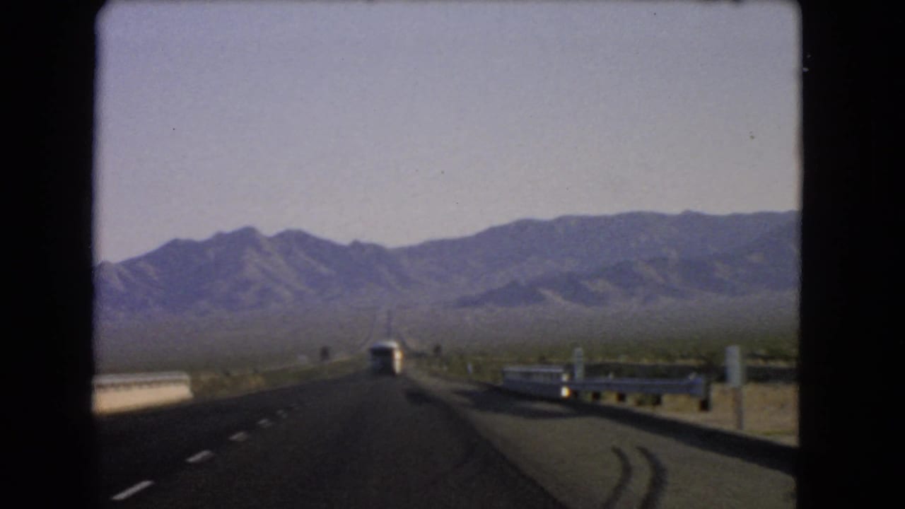 1966 Nevada home-movie POV from a moving car, looking past guardrails and a bus toward a hazy mountain range.