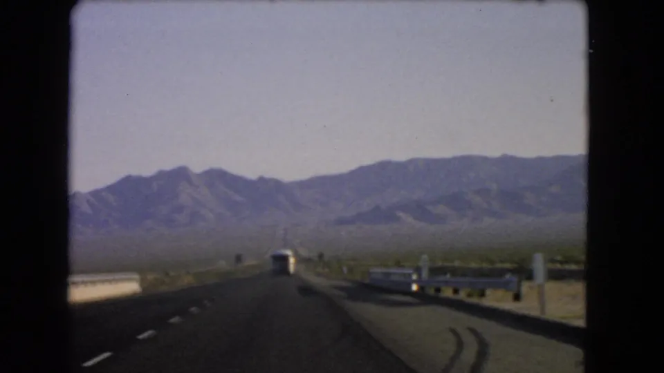 1966 Nevada home-movie POV from a moving car, looking past guardrails and a bus toward a hazy mountain range.