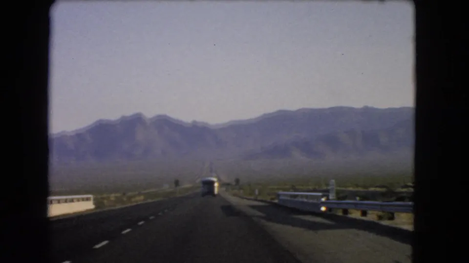 1966 Nevada road-trip footage showing a straight highway, guardrail, white bus, desert scrub, and distant mountains from a car windshield view.