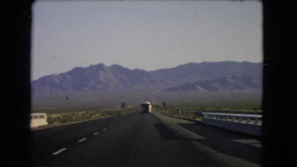 Moving-car view of a Nevada highway in 1966, with guardrails, a white bus, open desert terrain, and a mountain range beyond.