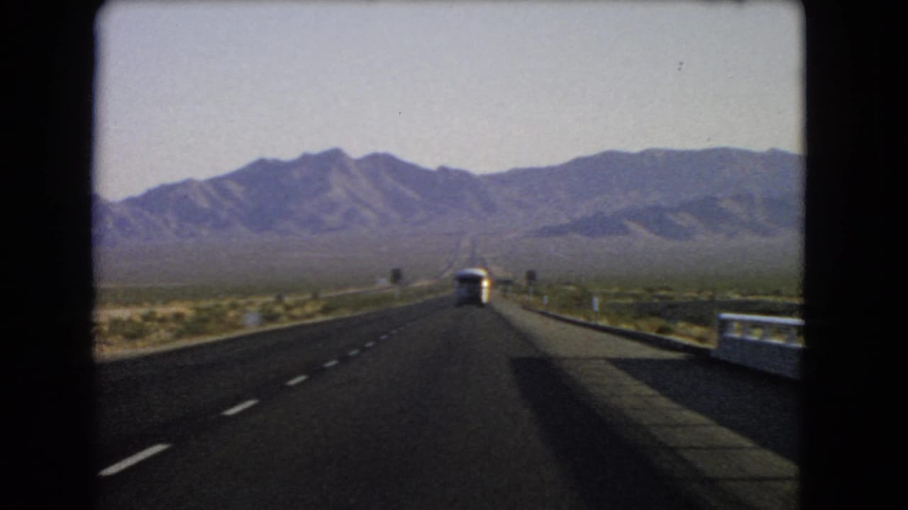 1966 Nevada home-movie frame from a car on a desert highway, showing a bus ahead, road signs, guardrail, scrubland, and mountains.