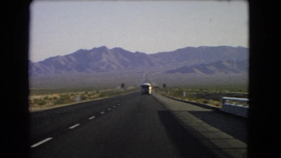 1966 Nevada home-movie frame from a car on a desert highway, showing a bus ahead, road signs, guardrail, scrubland, and mountains.