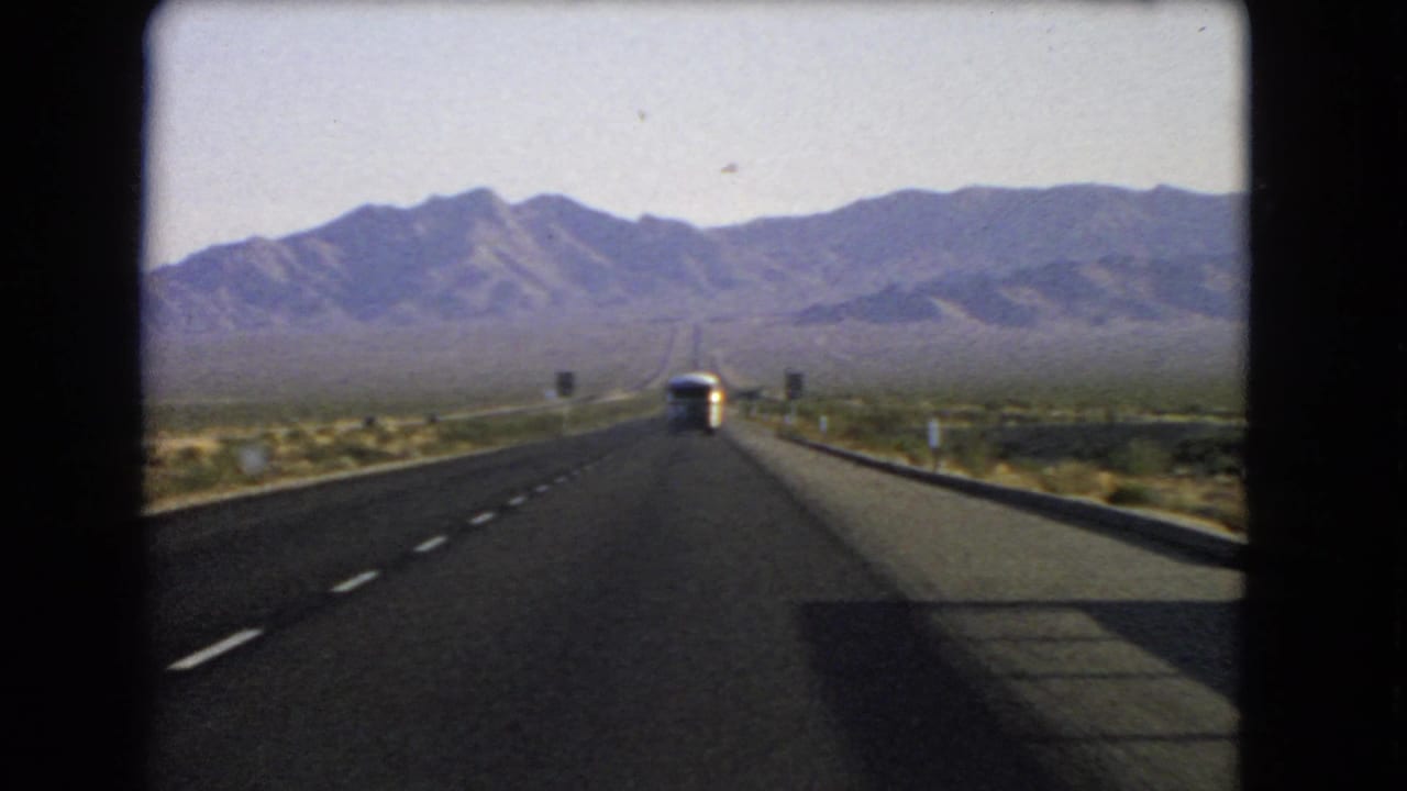 Final 1966 Nevada highway frame showing the moving car perspective, bus ahead, low barriers, desert brush, and mountain backdrop.