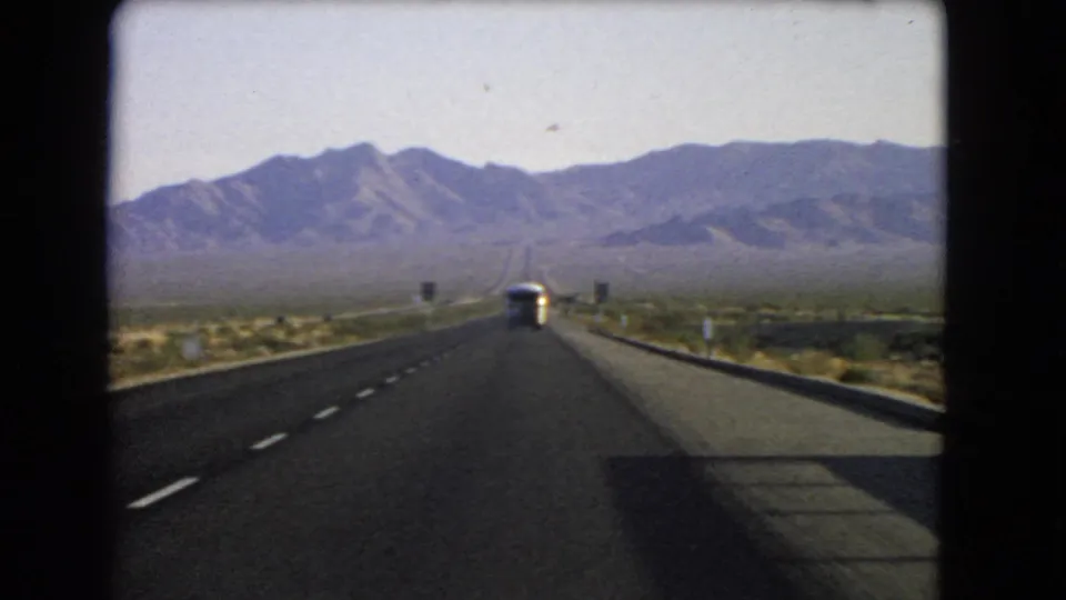Final 1966 Nevada highway frame showing the moving car perspective, bus ahead, low barriers, desert brush, and mountain backdrop.