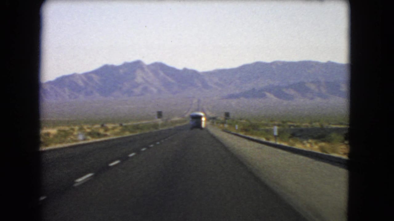 1966 Nevada 8mm road-trip view down a straight desert highway toward distant rocky mountains with a bus ahead.