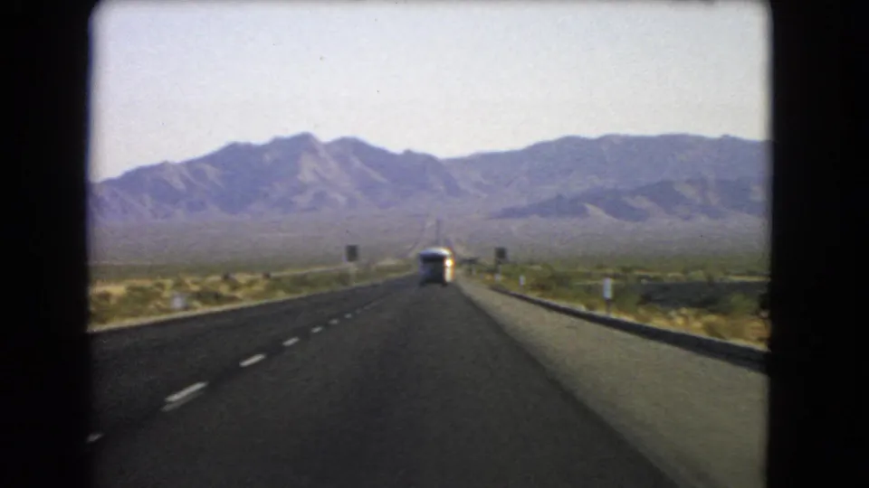 1966 Nevada 8mm road-trip view down a straight desert highway toward distant rocky mountains with a bus ahead.