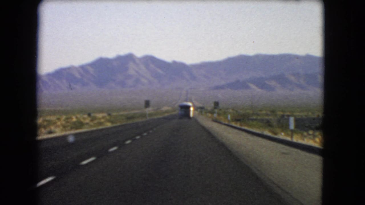 1966 Nevada home-movie frame showing a highway stretching through arid desert toward mountains, with a bus in the lane ahead.