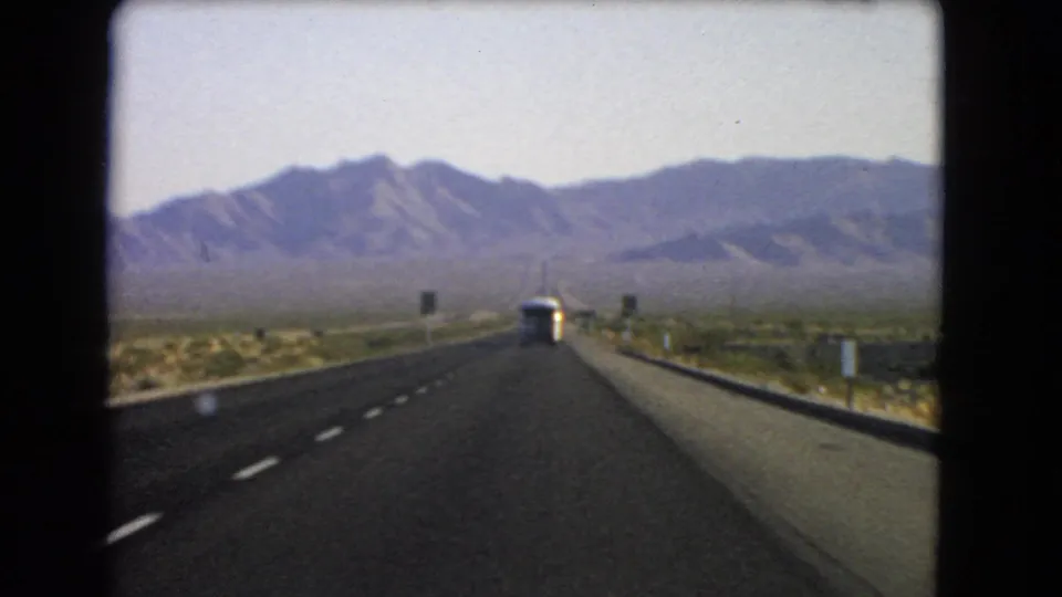1966 Nevada home-movie frame showing a highway stretching through arid desert toward mountains, with a bus in the lane ahead.