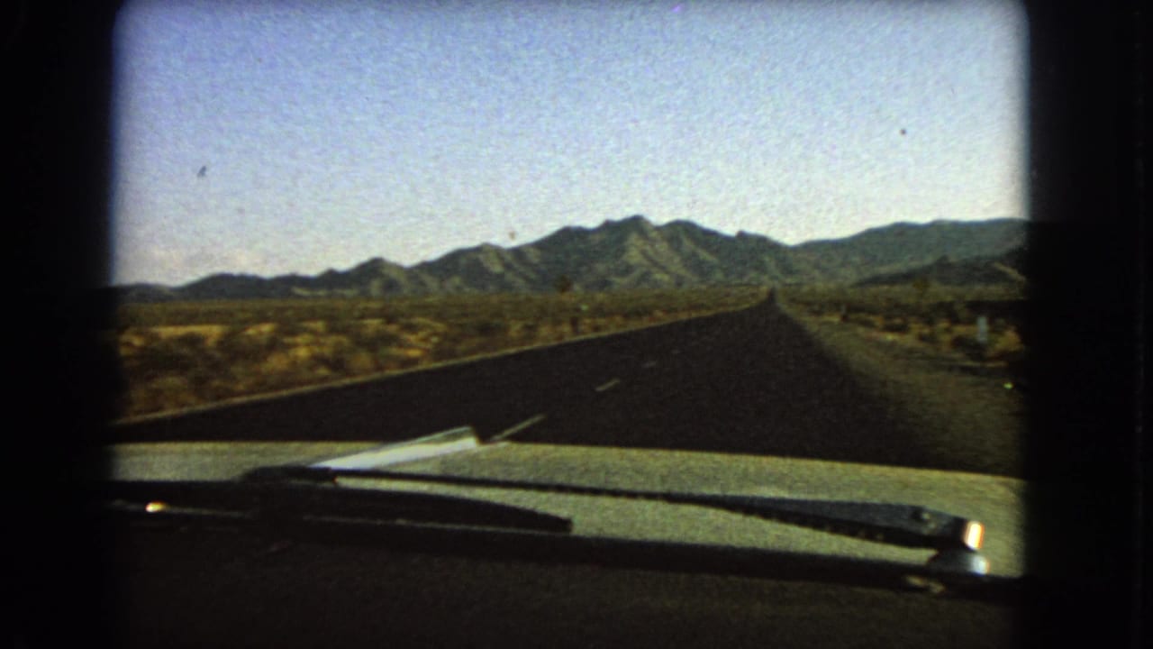 1966 Nevada 8mm view from inside a moving car, looking past the hood and wiper toward a straight desert road and mountains.