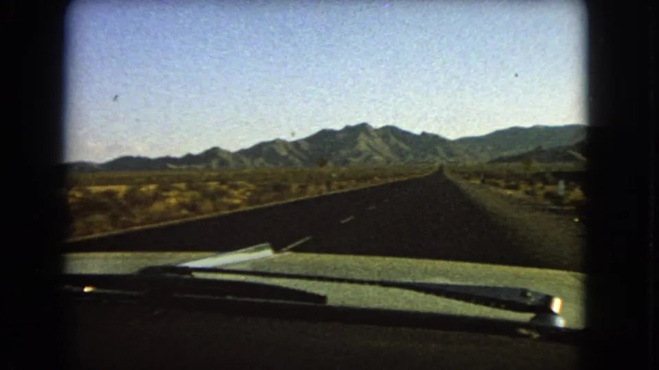 1966 Nevada 8mm view from inside a moving car, looking past the hood and wiper toward a straight desert road and mountains.