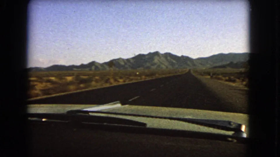 1966 Nevada home-movie frame showing a forward car view on a straight desert highway with mountains on the horizon.