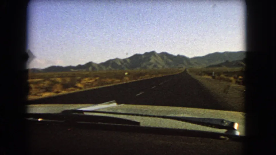 Passenger-seat 8mm frame from Nevada in 1966, with the car hood at bottom and a straight road crossing an arid plain.