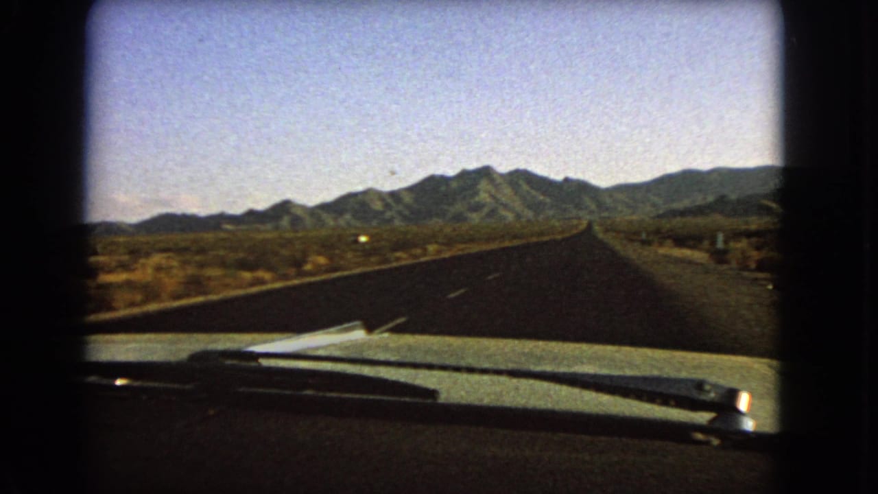 1966 Nevada 8mm travel frame looking over the windshield area toward a straight desert highway and distant mountain range.