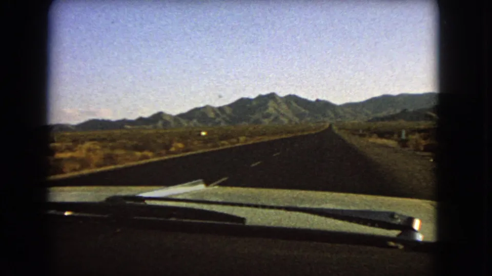 1966 Nevada 8mm travel frame looking over the windshield area toward a straight desert highway and distant mountain range.