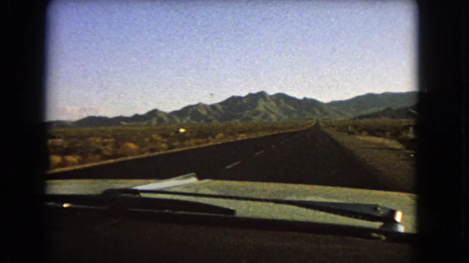 1966 8mm Nevada road view over a car hood, with windshield wipers, desert terrain, and distant mountains under a clear sky.