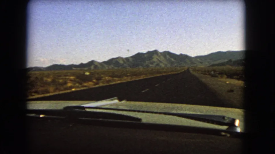 First-person 8mm view from a car on a Nevada desert highway, with the road running toward low mountains and open scrubland.
