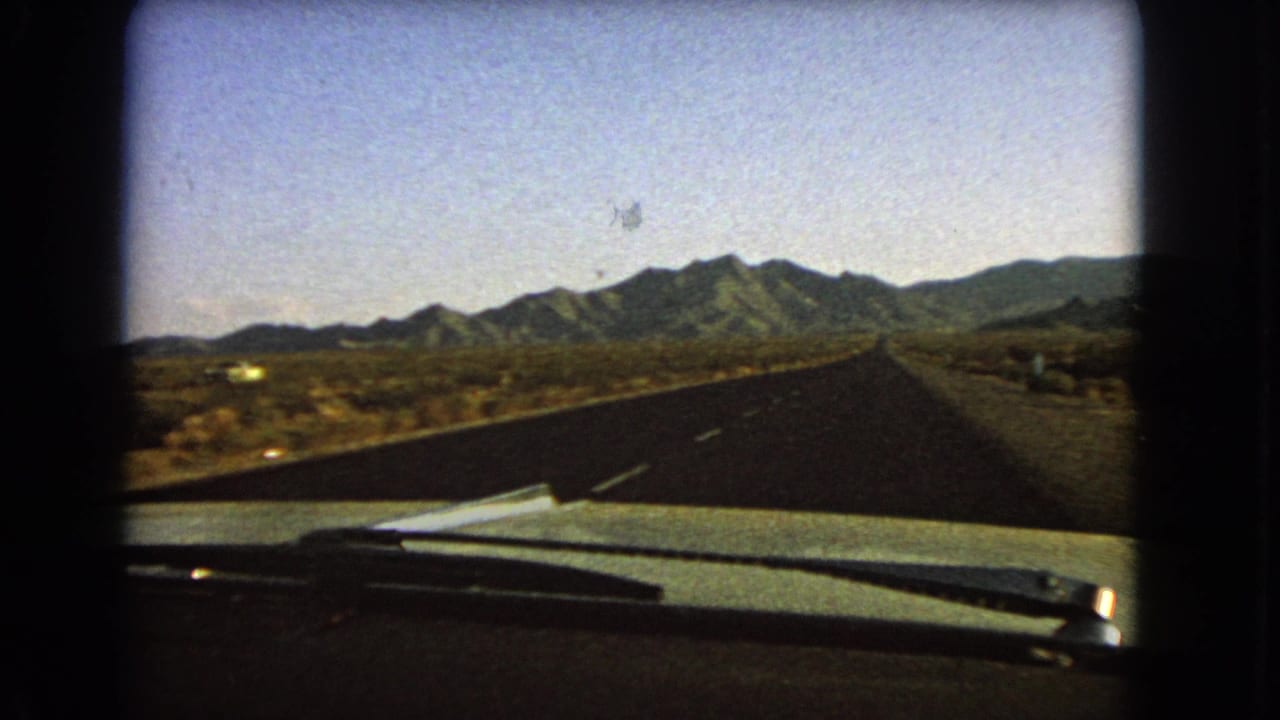 Vintage 8mm Nevada driving view showing an empty paved highway, dashboard edge, windshield wipers, desert brush, and mountains ahead.