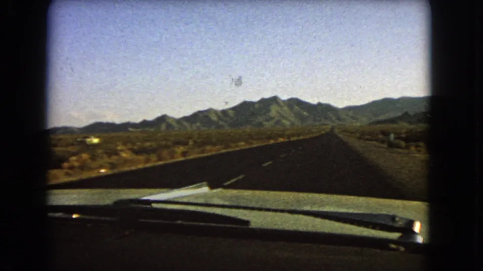Vintage 8mm Nevada driving view showing an empty paved highway, dashboard edge, windshield wipers, desert brush, and mountains ahead.