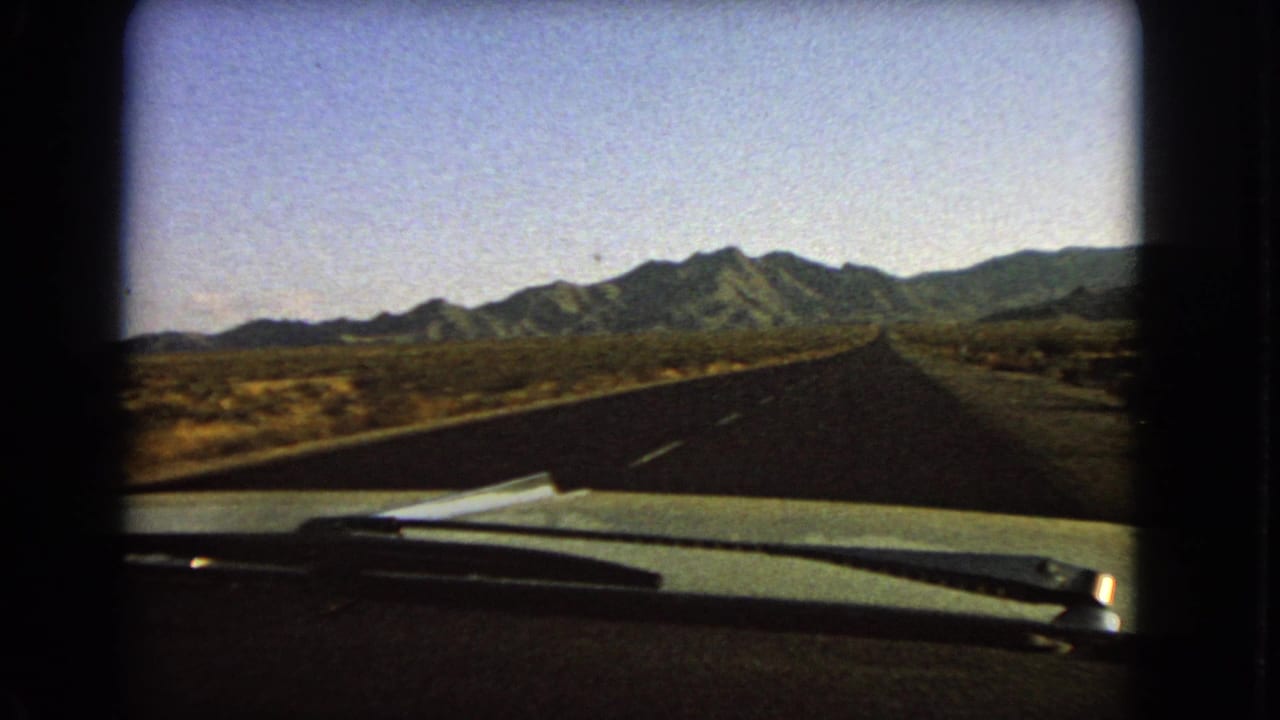 Late frame from a 1966 Nevada 8mm road sequence, showing a car hood, windshield wipers, a quiet highway, desert scrub, and distant mountains.