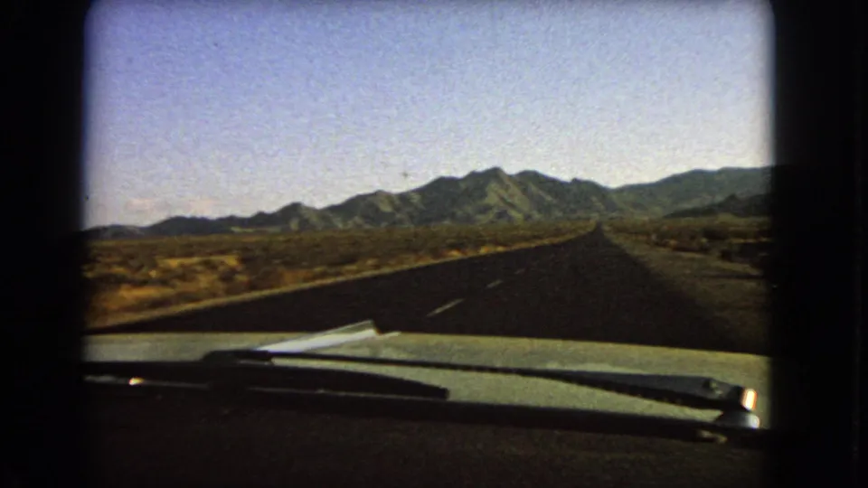 Late frame from a 1966 Nevada 8mm road sequence, showing a car hood, windshield wipers, a quiet highway, desert scrub, and distant mountains.