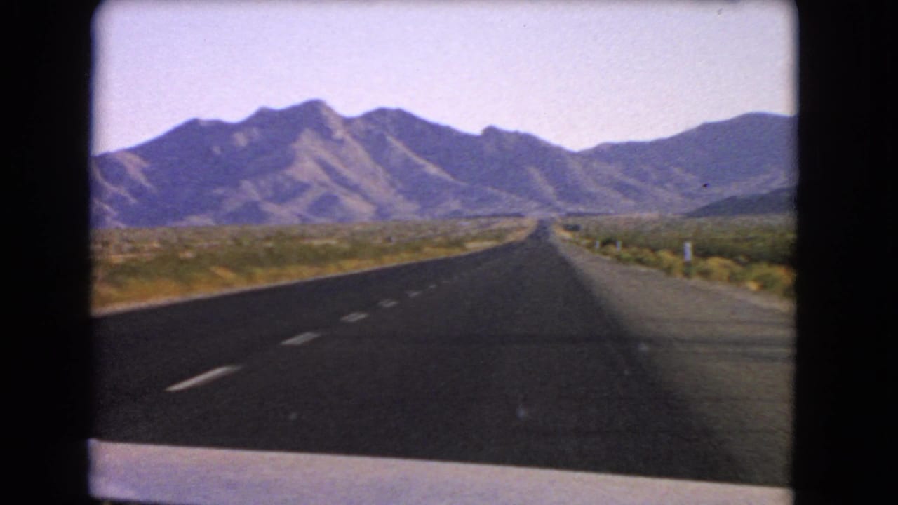 1966 Nevada 8mm frame from inside a moving vehicle, looking down a highway toward rugged desert hills.