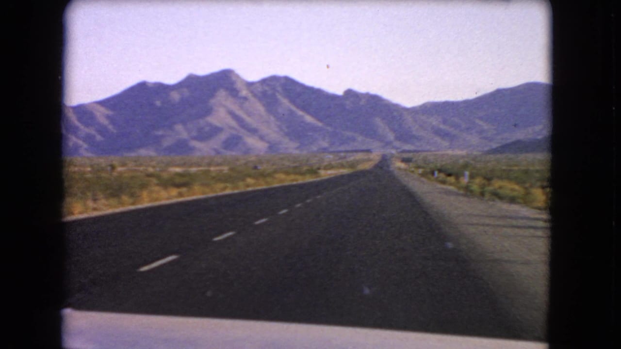 1966 Nevada home-movie frame showing a highway from inside a moving vehicle, with desert scrub and mountains ahead.