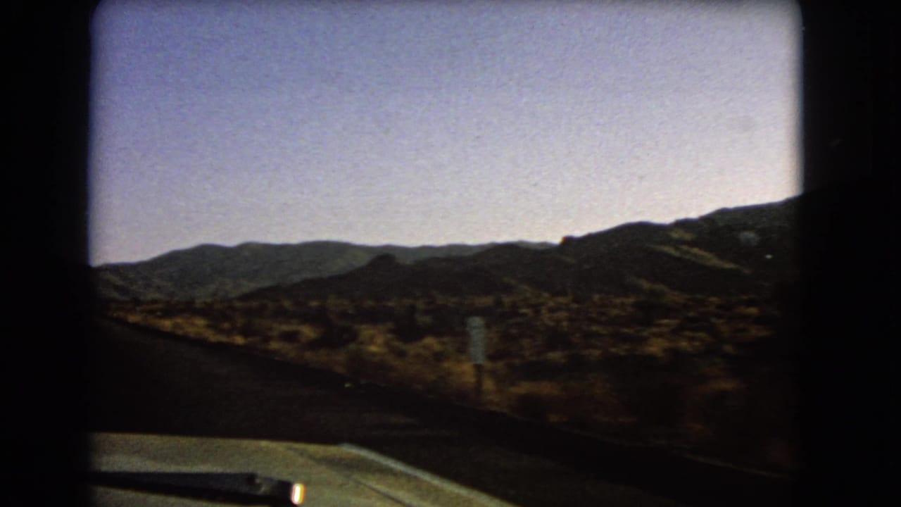 Moving-car view across Nevada desert terrain, with the dashboard at the bottom and low mountains on the horizon.