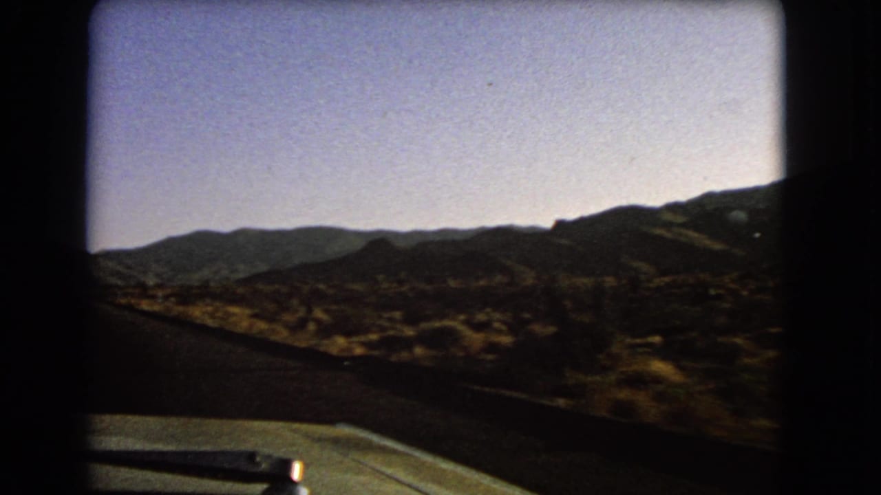 1966 Nevada 8mm frame from a moving car, looking past the dashboard toward scrubland and low mountain ridges.