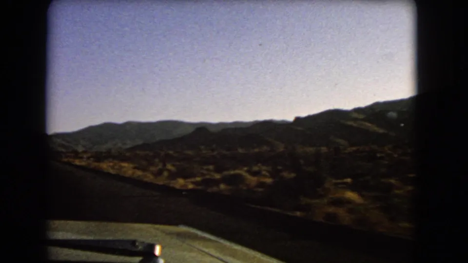 1966 Nevada 8mm frame from a moving car, looking past the dashboard toward scrubland and low mountain ridges.