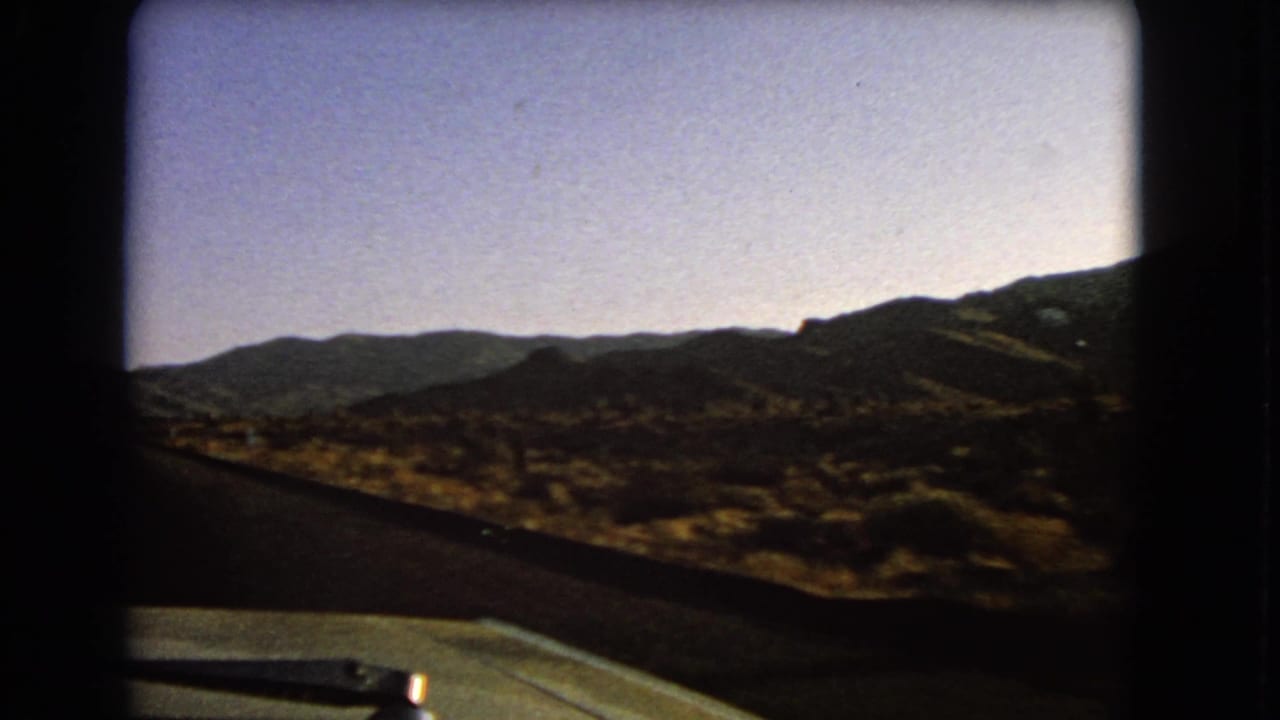 1966 Nevada home-movie view from inside a car, with the hood, wiper, asphalt road, dry brush, and distant ridges visible.