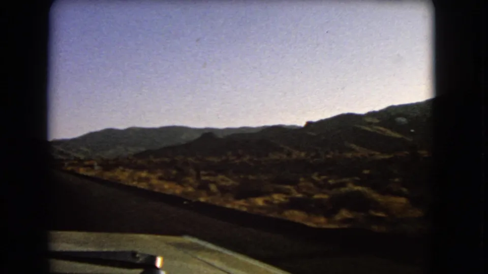 1966 Nevada home-movie view from inside a car, with the hood, wiper, asphalt road, dry brush, and distant ridges visible.
