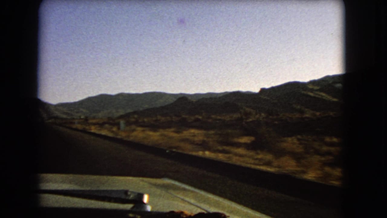 1966 Nevada 8mm frame showing the road from a moving car, with desert scrub, low mountains, windshield edge, and car hood in view.