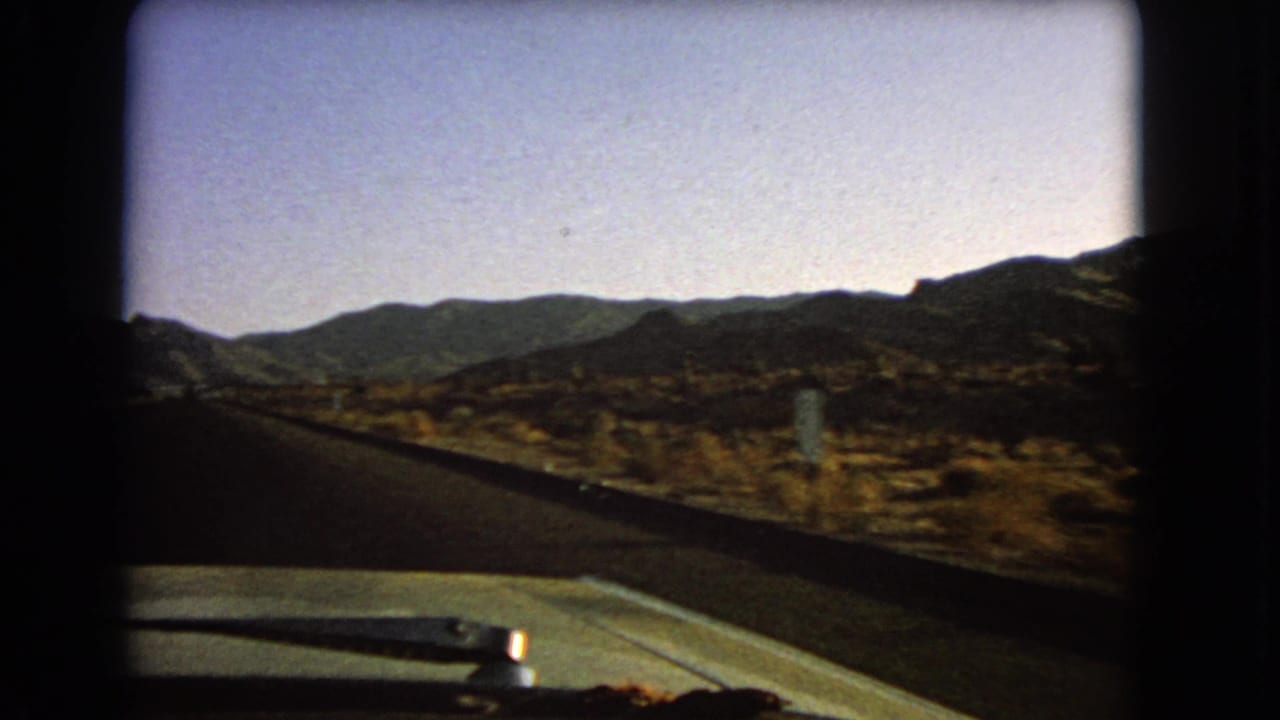 Passenger-side 8mm view in Nevada, 1966, showing a dark road, pale hood, roadside desert brush, and layered hills in the distance.